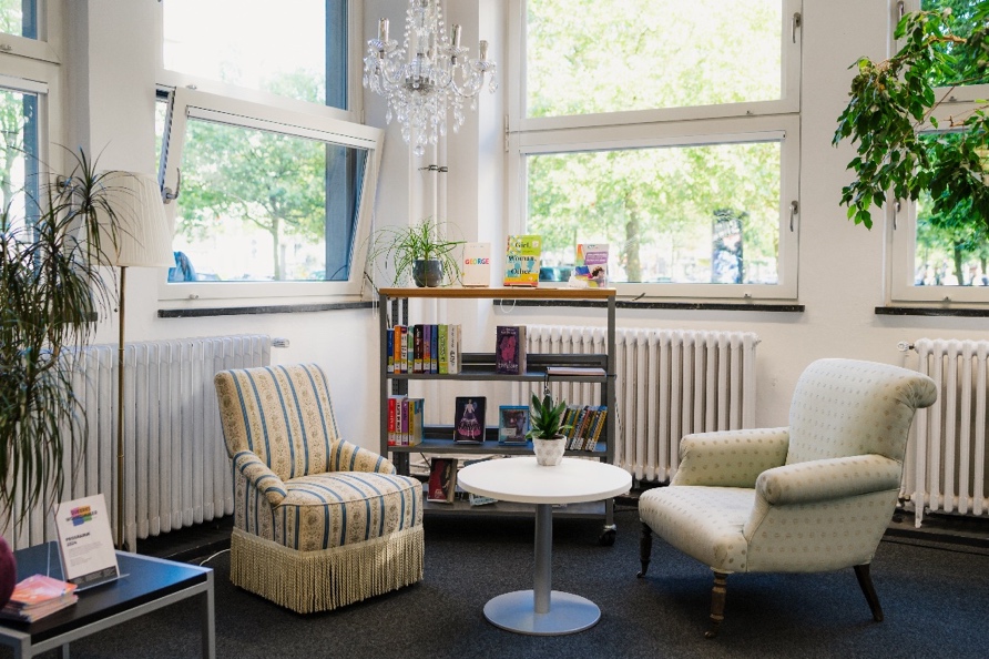 <i>Die Sitzecke im Queeren Wohnzimmer. Foto: Olli Lehne</i> Zwei Sessel, ein Bücherregal, ein Tischchen und ein Kronleuchter umgeben von Pflanzen.