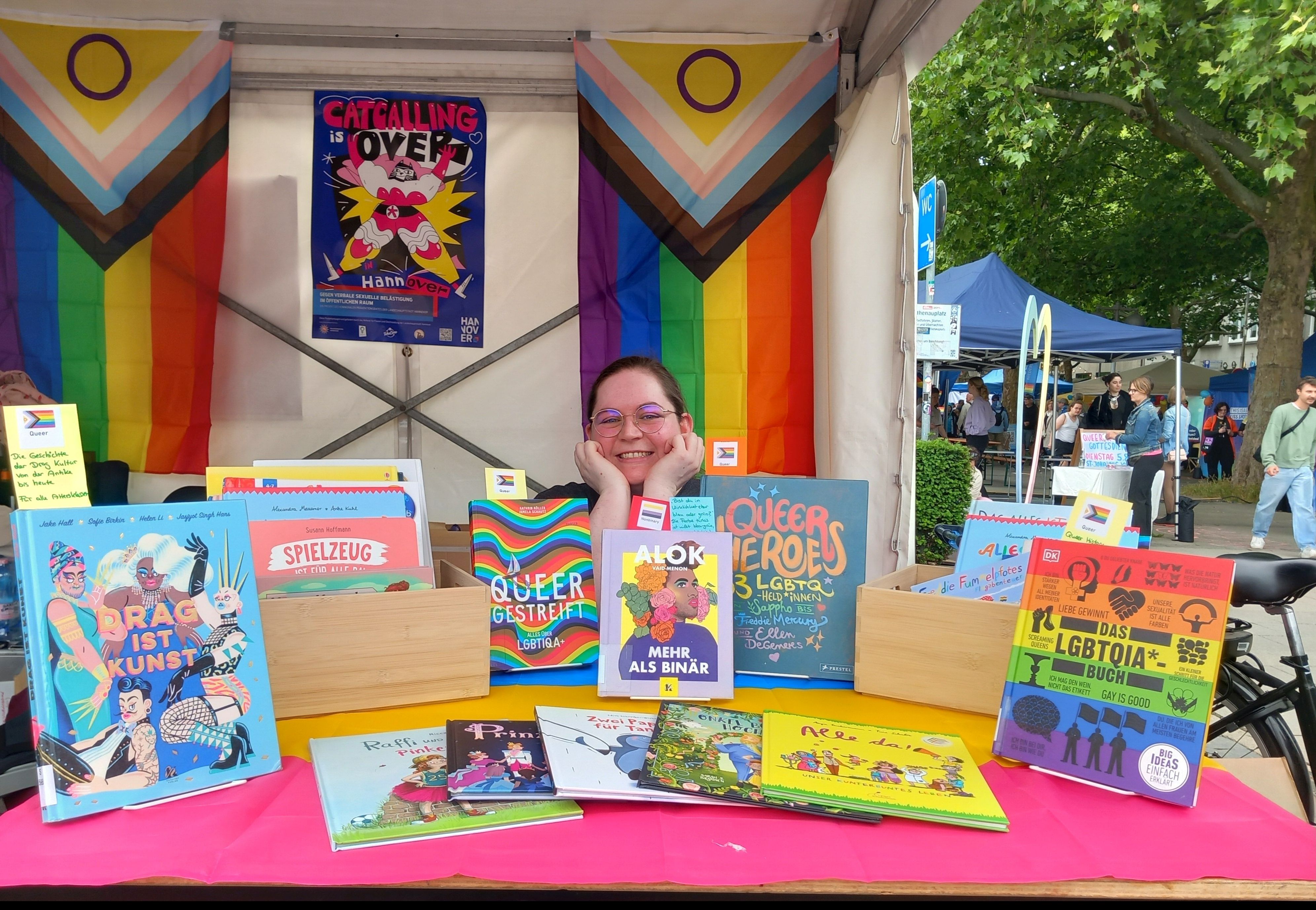 <i>Der Stand der Stadtbibliothek Hannover auf dem CSD; Foto: Ramona Brase</i> Stand mit regenbogenfarbenen Büchern mit zwei Prideflaggen im Hintergrund.