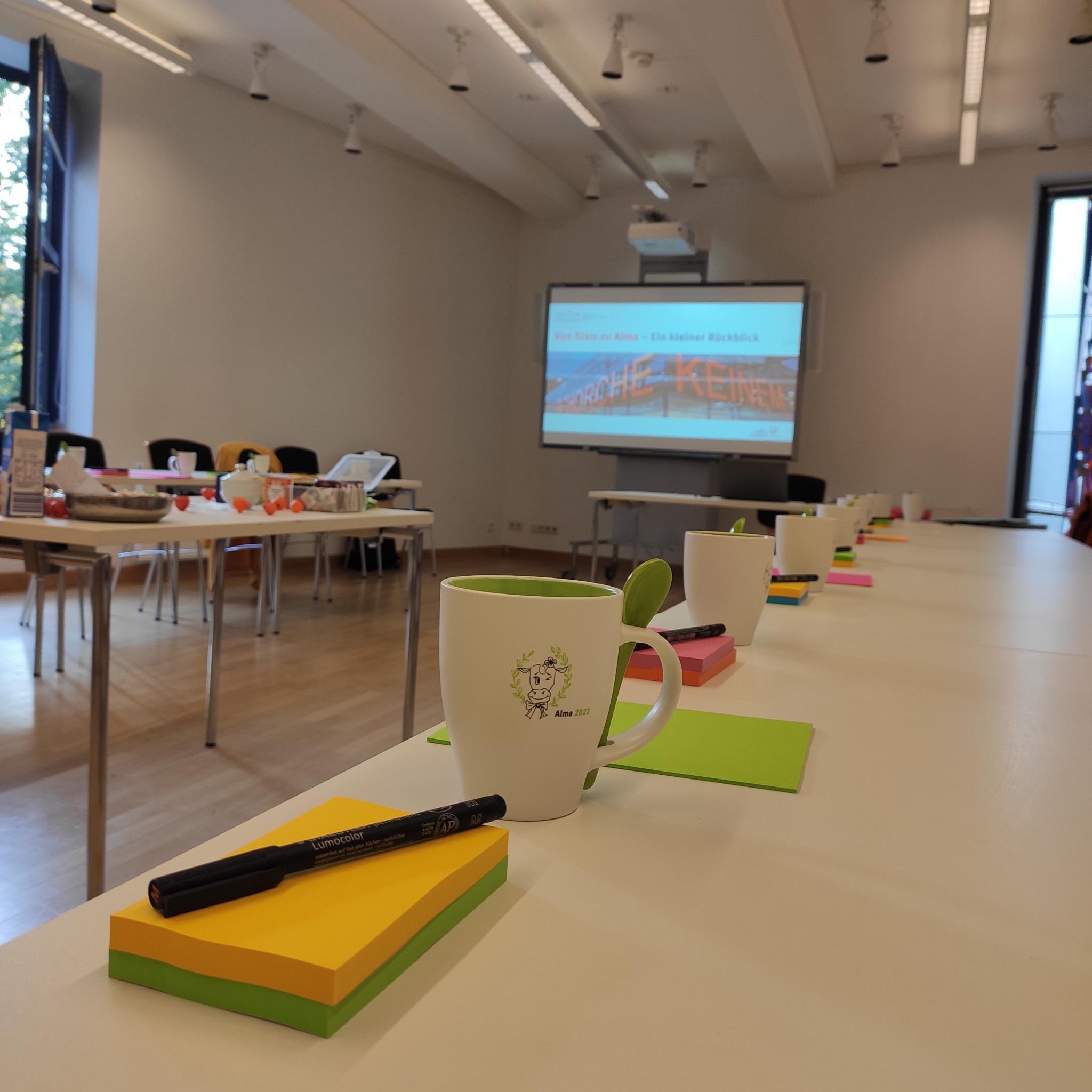 <i>Kurz vor Beginn der Abschlussbesprechung</i> Ein Foto eines Besprechungsraum. Im Vordergrund sind bunte Notizzettelblöcke und Stifte sowie Becher zu sehen, die mit dem Alma-Kuh-Maskottchen verziert sind; im Hintergrund wird die Titelseite einer Präsentation auf einer Beamer-Projektionsfläche gezeigt.