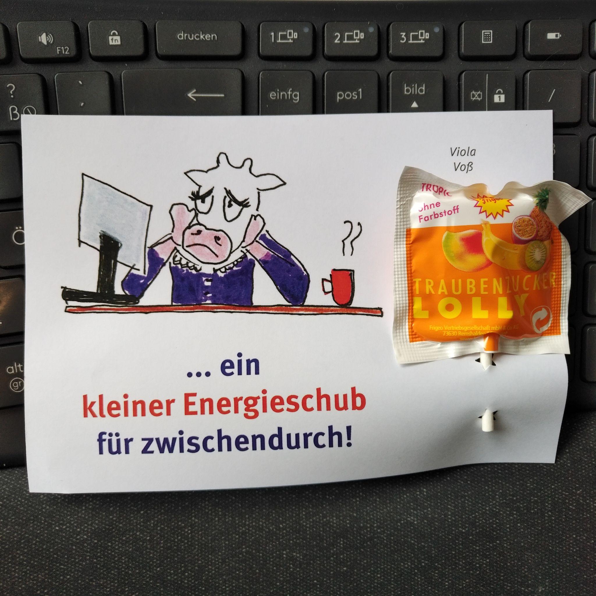 <i>Es gab auch eine Zeichnung des Maskottchens, das müde am Schreibtisch sitzt – ein Aufhänger für einen kleinen “Doping-Gruß”.</i> Ein Foto, das einen bunt ausgedruckten Zettel auf einer Tastatur liegend zeigt. Darauf ist eine Zeichnung mit einer verzweifelt dreinschauenden Kuh in einer lilafarbenen Bluse zu sehen. Sie schaut auf einen Bildschirm; neben ihr steht eine dampfende Tasse Kaffee. Unter der Zeichnung steht der Satz "… ein kleiner Energieschub für zwischendurch!", der sich auf den Traubenzucker-Lolli bezieht, der am rechten Teil des Zettels befestigt ist.