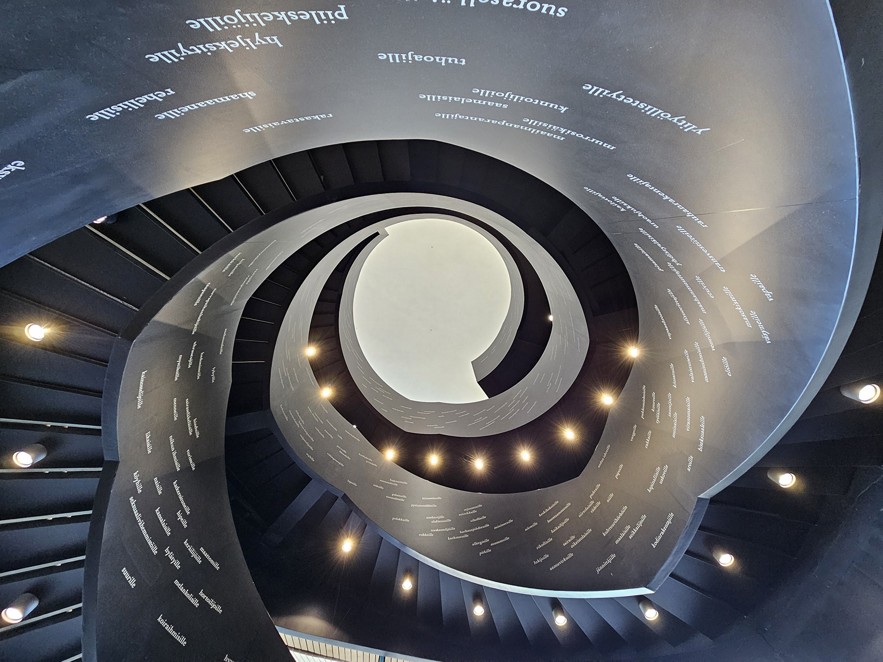Wie eine Spirale zieht sich die Innentreppe der Oodi-Bibliothek nach oben. Auf den schwarzen Wänden sind Aufschriften in Weiß zu sehen. 