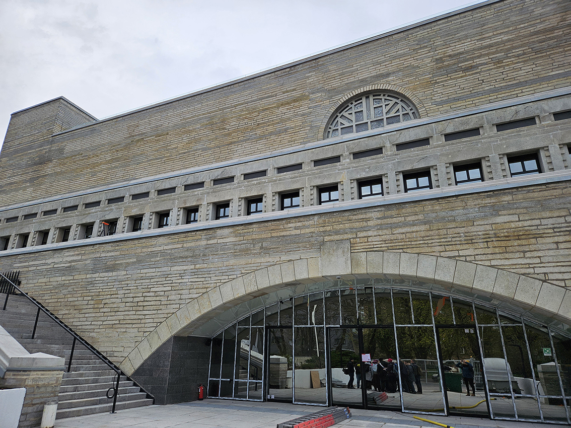 Auf der Abbildung ist der Eingangsbereich der Nationalbibliothek Tallinn mit einem geschwungenen Türbereich zu sehen. Links führt eine große Treppe nach oben. 