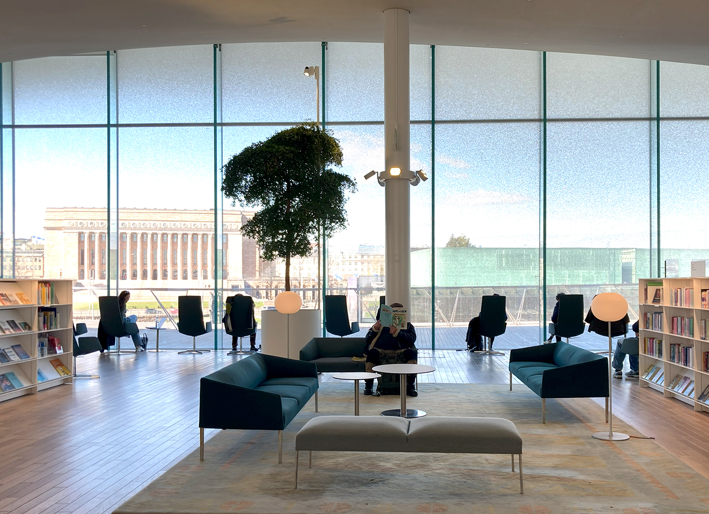 Das Bild zeigt im Vordergrund den Lesebereich im Obergeschoss der Oodi-Bibliothek. Durch die große Glasfassade geht der Blick nach außen auf den Bürgerplatz von Helsinki. 