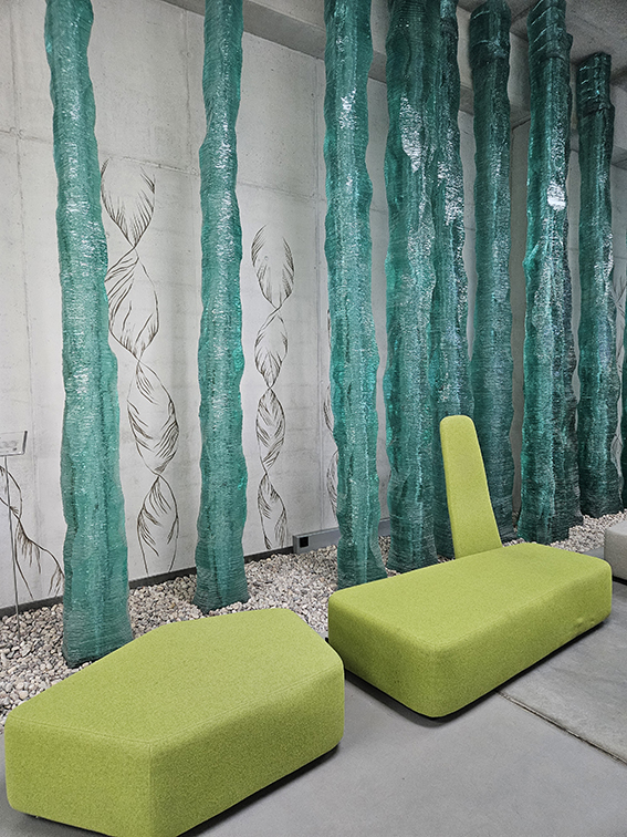 Es sind zwei grüne Sitzbänke zu sehen. Sie stehen vor acht Säulen aus grünlichem Glas und einer Wand, auf der sich Zeichnungen von Helixstrukturen befinden.  