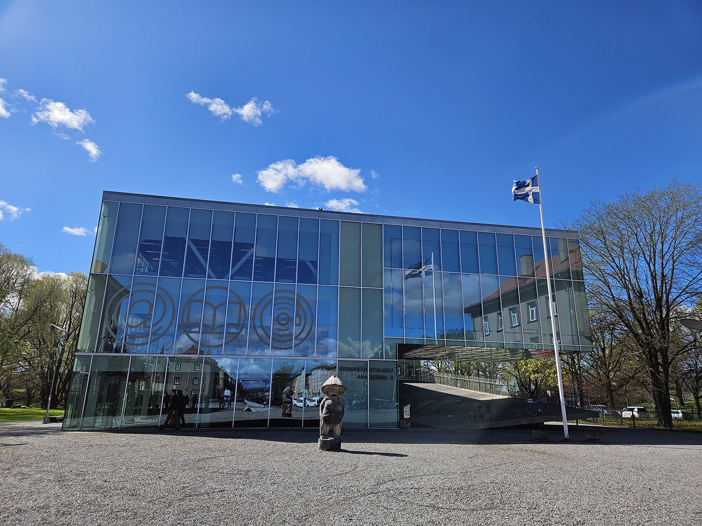 Die Abbildung zeigt die Stadtbibliothek Pärnu mit einem kleinen Denkmal vor dem Eingang und der Spiegelung des Stadtraums in der Glasfassade. 