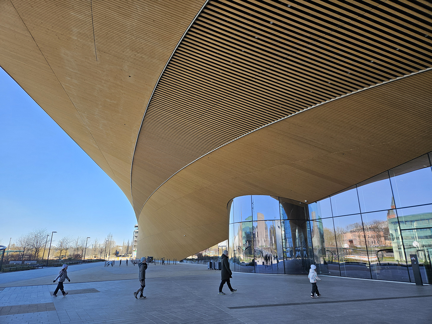 Auf dem Bild ist der Außenbereich vor dem Eingang der Oodi-Bibliothek mit dem auffälligen, weit auskragenden Dach zu sehen. Vier Menschen laufen auf die Eingangstür zu. 