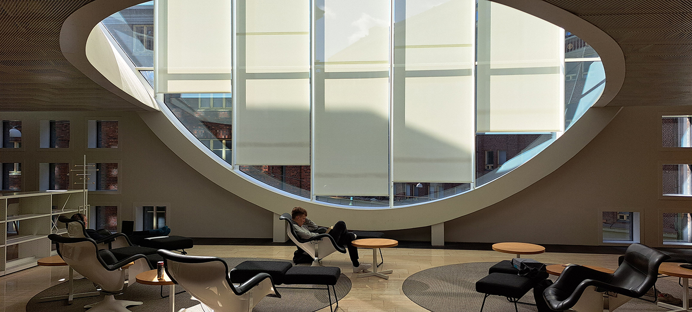 Ein junger Mann sitzt entspannt vor einem großen runden Fenster im Loungebereich des Lesesaals des Kaisa House Helsinki.