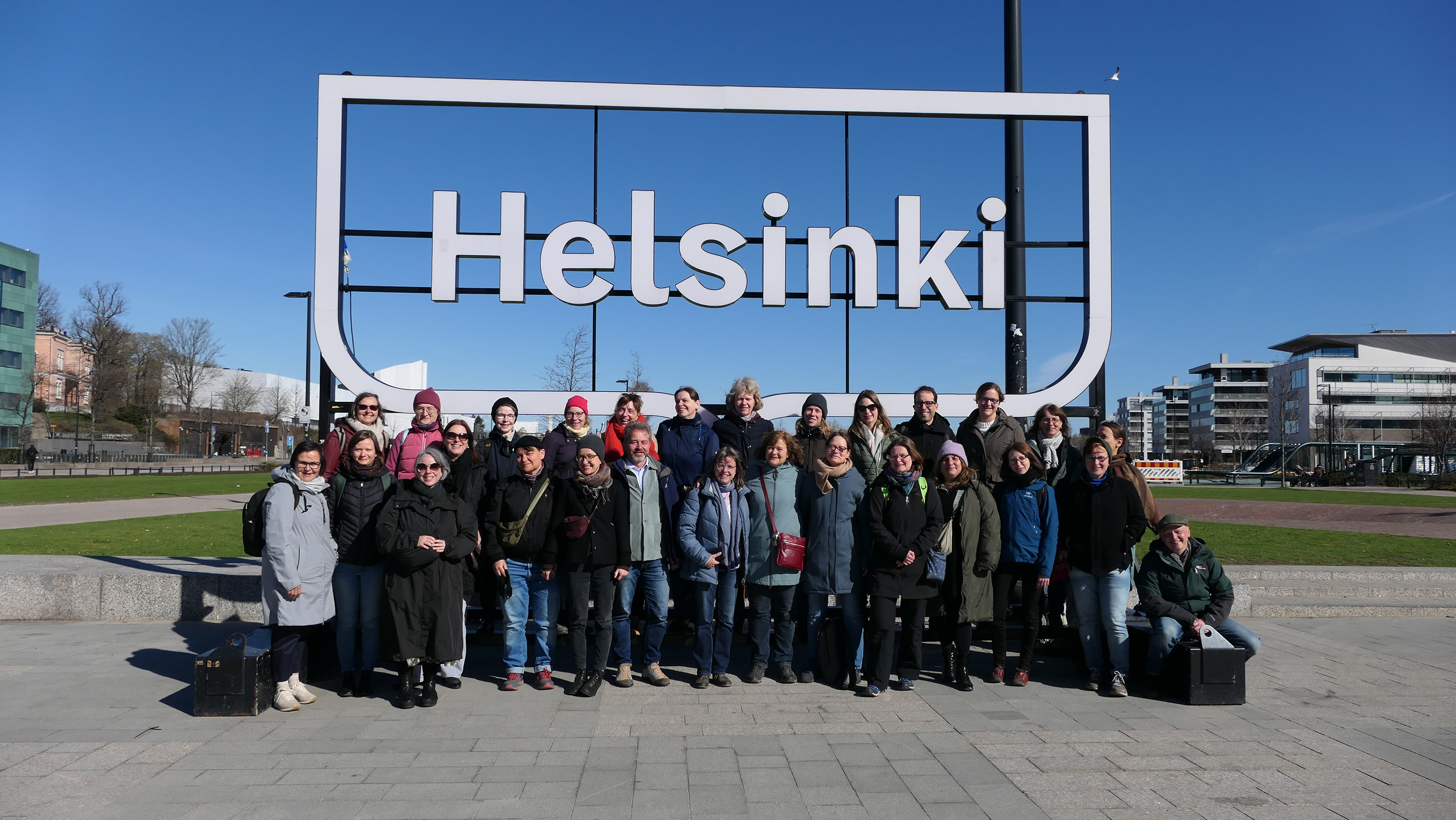 Das Bild zeigt die Reisegruppe der Exkursion vor blauem Himmel und dem Schriftzug "Helsinki". 