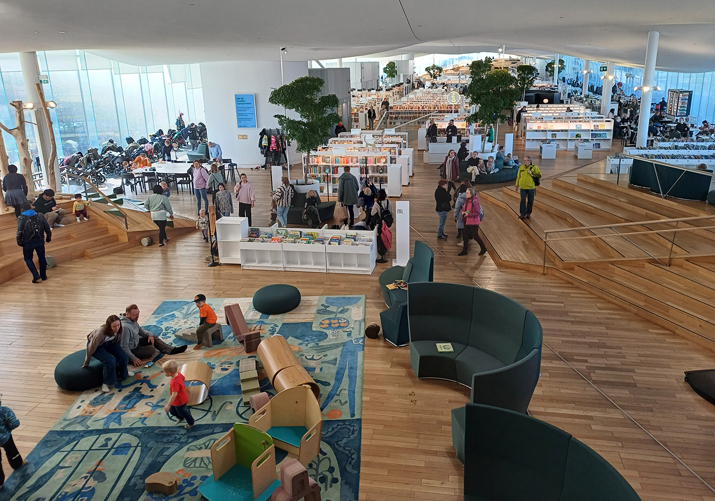 Im Kinderbereich der Oodi-Bibliothek spielen im Vordergrund des Bildes Familien auf einem Teppich. Im Hintergrund erstreckt sich der Bereich des sogenannten "Bücherhimmels". 