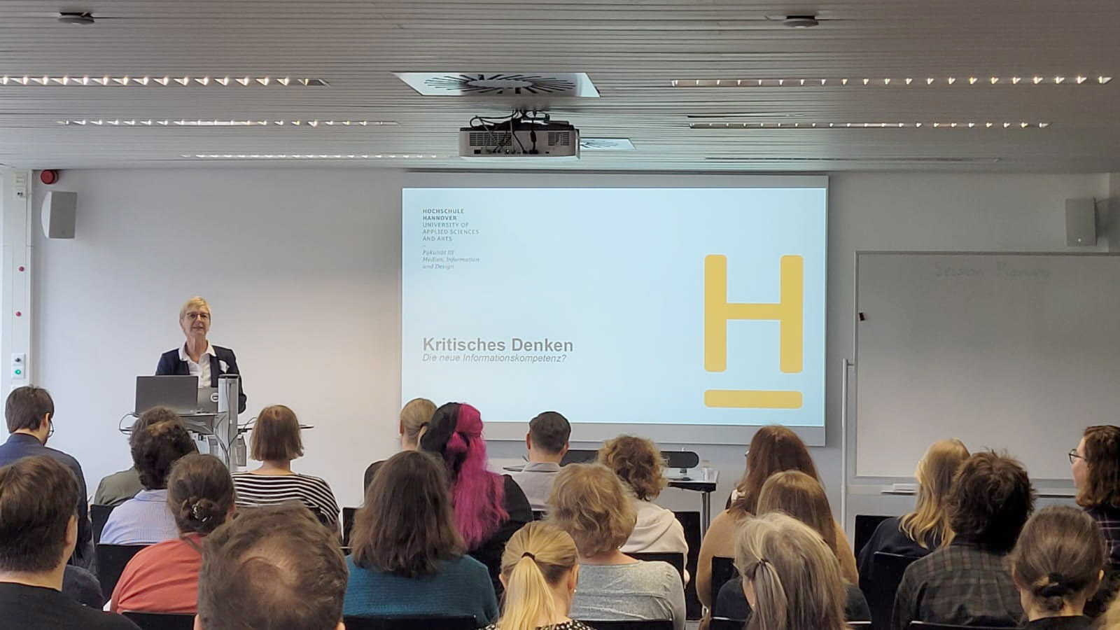 <i>Anke Wittich präsentiert ihre Keynote, Foto: TIB / L. Gericke</i> Anke Wittich während ihrer Keynote vor den Teilnehmenden, die von hinten zu sehen sind. Hinter ihr ist die Titelfolie ihrer Präsentation an die Wand projiziert.