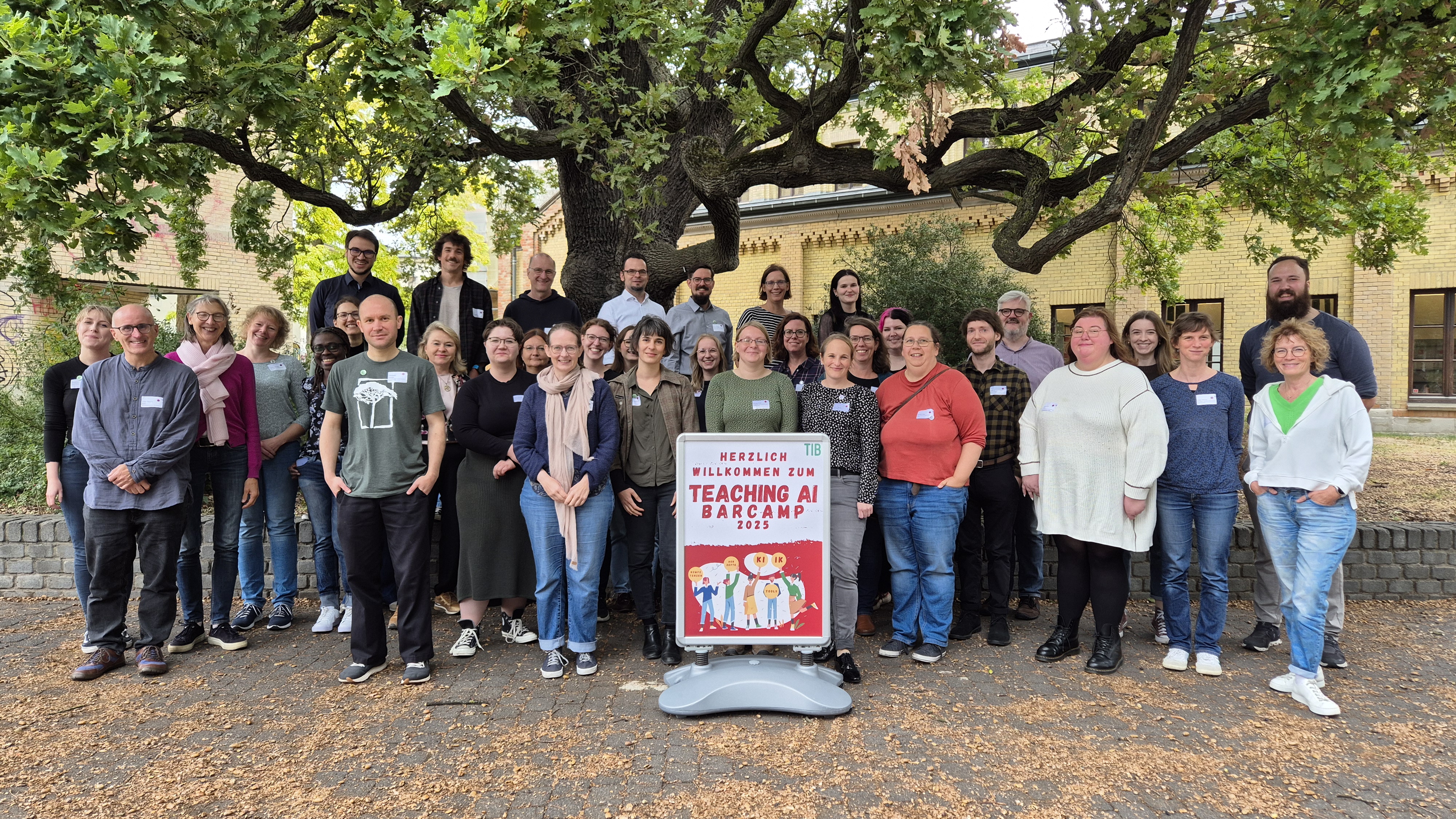 <i>Gruppenbild der Teilnehmenden, Foto: TIB / A. Bätje</i> Gruppenfoto aller Teilnehmenden auf dem Platz vor dem Hauptgebäude der TIB Hannover. Im Vordergrund ist ein Veranstaltungsplakat des Barcamps zu sehen. Im Hintergrund ist das Marstallgebäude der TIB Hannover.