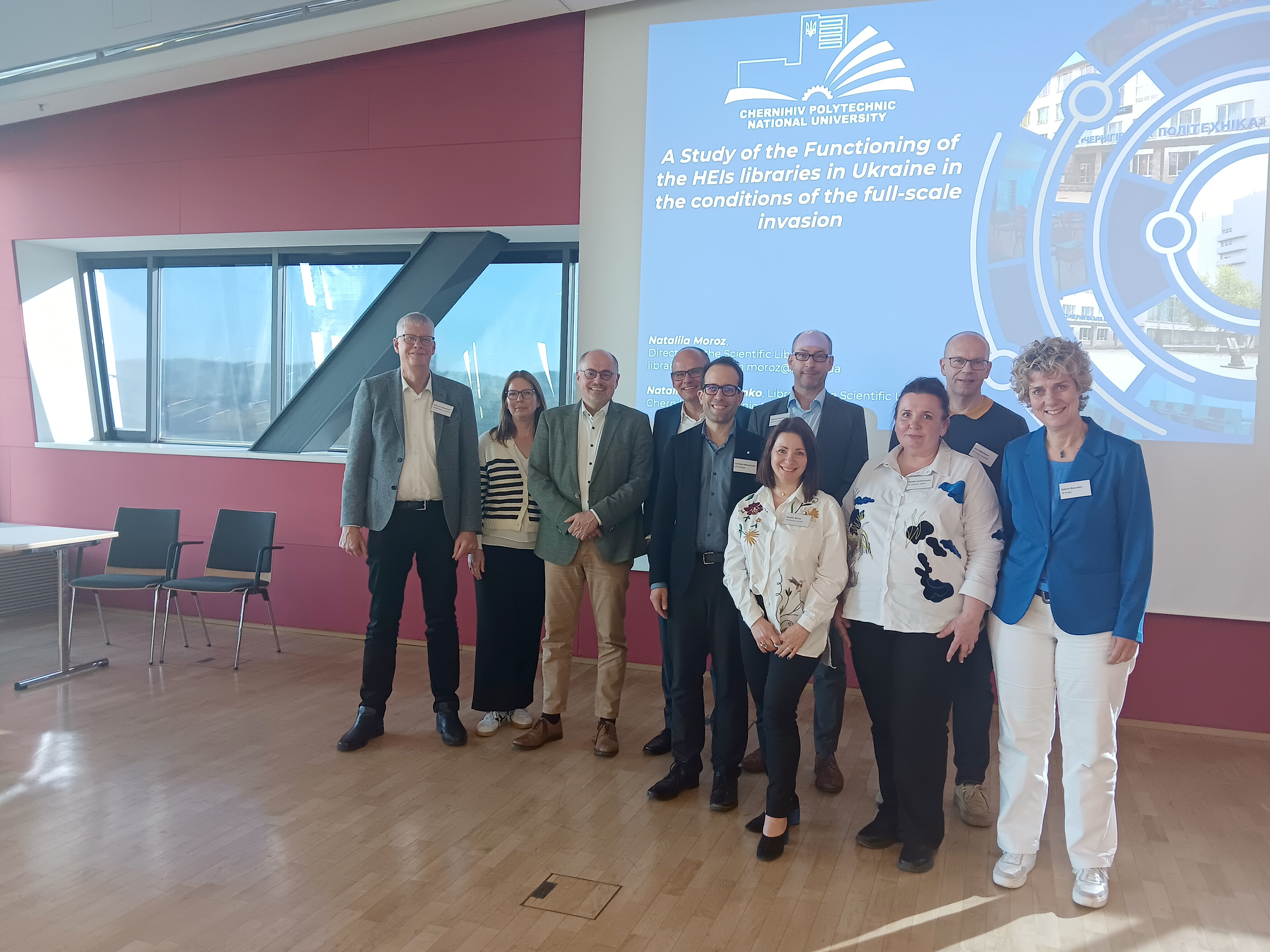 Eine Gruppe von Menschen steht in einem Konferenzraum vor einer Leinwand. Die Leinwand zeigt eine Präsentationsfolie mit einem blauen Hintergrund, mehreren Logos und einem englischen Titel. Die Personen tragen überwiegend formelle Kleidung wie Hemden, Blazer und Blusen. Der Raum ist hell und modern, mit großen Fenstern und Holzfußboden. Im Hintergrund sind einige Stühle und Tische zu sehen.