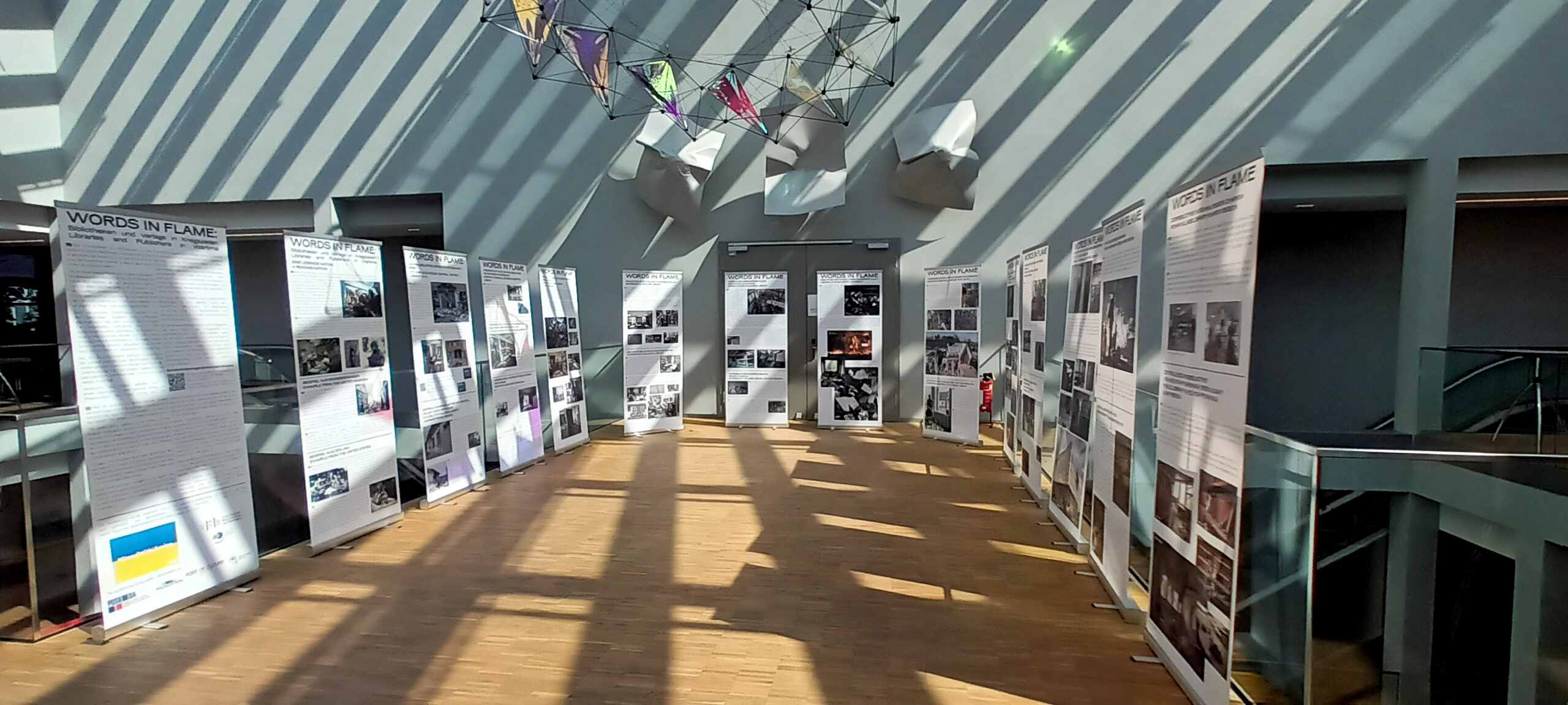 In einem lichtdurchfluteten Raum mit diagonalen Schattenstreifen auf dem Boden und den Wänden sind mehrere große, aufrecht stehende Infotafeln zu sehen. Die Tafeln zeigen zahlreiche Fotografien und Textblöcke. Im Hintergrund hängen geometrische, bunte Dekorationen von der Decke. Der Boden besteht aus hellen Holzplanken, und das Geländer aus Glas und Metall begrenzt den Raum.